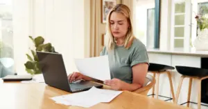 A blond woman ordering a report online while looking at a paper. 