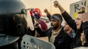 A group of protestors confronting an officer in riot gear and a shield. 